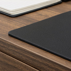 Desky black leather desk mat with pebbled texture on a wooden desk, notebook in background.